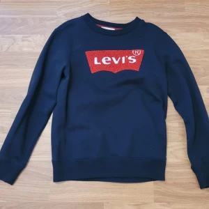 Svart sweatshirt från Levi's - Svart sweatshirt från Levi's med klassisk röd logga framtill. Tröjan har rund halsringning och långa ärmar. Perfekt för en avslappnad stil.