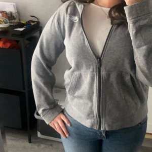 Grå zip-hoodie från Nike - Grå hoodie från Nike med dragkedja framtill och två fickor. Klassisk vit Nike-logga på bröstet. Mjuk och bekväm, perfekt till vardags. Modellen är enkel och sportig med lång ärm och huva.