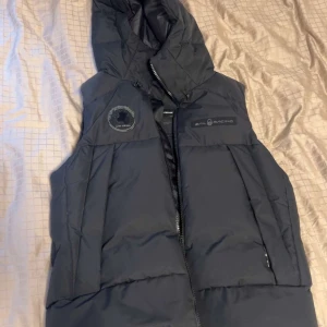 Svart dunväst med huva från Sail Racing - Svart dunväst från Sail Racing med stor huva och dragkedja framtill. Västen har två sidofickor med dragkedja och logotyp på bröstet. Perfekt för lager-på-lager och håller dig varm under kyliga dagar.