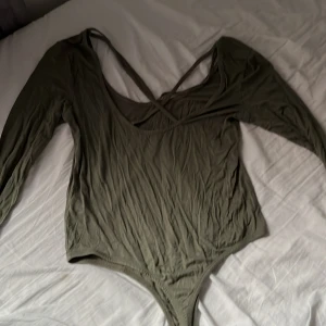 Olivgrön långärmad body - Säljer en olivgrön body med långa ärmar och djup ringning framtill. Bodyn har smala axelband som korsas i ryggen för en snygg detalj. Perfekt att styla med jeans eller kjol.