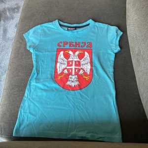 Turkos t-shirt med Serbiens vapen från 011 - Turkos t-shirt från 011 med stort tryck av Serbiens vapen och texten 'Srbija' på bröstet. Klassisk rund hals och korta ärmar. Perfekt för dig som vill visa ditt stöd för Serbien eller gillar unika tryck.