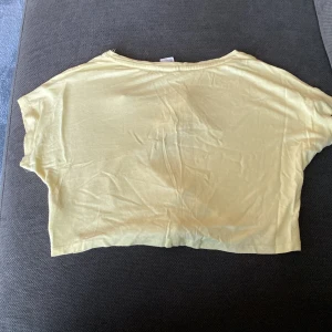 Gul croppad t-shirt med tukan och paljetter från H&M - Säljer en ljusgul croppad t-shirt från H&M x Kate Morgan Studio med tryck av en tukan, texten 'All you need is summer!' och färgglada paljetter på framsidan. Toppen har korta ärmar och knytning nedtill.