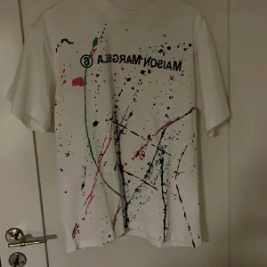 Vit t-shirt med färgstänk från Maison Margiela - Vit t-shirt från Maison Margiela med färgstänk i grönt, rött, svart och lila samt tryckt logga framtill. Klassisk passform och rund halsringning. Perfekt statement-plagg för dig som gillar unika designer.
