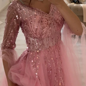 Rosa glittrig balklänning med paljetter och tyll - Säljer en rosa långklänning med glittriga paljetter och tyll. Klänningen har trekvartsärm och v-ringning, samt volangdetaljer i kjolen. Perfekt för bal eller festliga tillfällen.