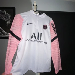 PSG långärmad träningslströja - Säljer en långärmad PSG fotbollströja i vitt med rosa ärmar och svarta detaljer. Tröjan har dragkedja vid halsen och tryck fram samt baktill. Den är helt ny, oanvänd, storlek S. 