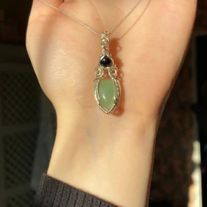 Halsband med grön sten och silverkedja - Unikt halsband med en grön oval sten infattad i detaljerad silvertråd och en liten svart sten ovanför. Hängs på en tunn silverkedja. Perfekt för dig som gillar naturinspirerade smycken och stilrena detaljer.