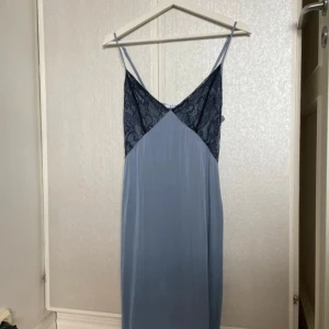 Ljusblå klänning med spetsdetaljer - Säljer en ljusblå långklänning med tunna axelband och v-ringning. Överdelen har svarta spetsdetaljer som ger en elegant touch. Klänningen har dragkedja i sidan och är tillverkad i ett mjukt, lätt material.