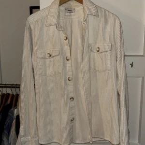 Vit ribbad skjorta/overshirt från Ciszere i manchestertyg - Säljer en vit ribbad skjorta/overshirt från Ciszere. Skjortan har långa ärmar, knappar framtill och två bröstfickor med lock. Broderad detalj nertill. Perfekt för dig som gillar stilrena plagg med lite struktur.