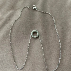 Silverfärgat halsband med cirkel och stenar - Säljer ett stilrent halsband i silverfärg med en tunn kedja och en rund cirkelberlock dekorerad med små gnistrande stenar runt kanten. Perfekt för dig som gillar minimalistiska smycken.