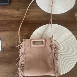 Ljusrosa axelväska med fransar från Maje - Ljusrosa axelväska från Maje i mocka❤️Väskan har både axelrem och handtag. Använt skick men fortfarande bra. Har lite fläckar på kanterna. Stora storleken