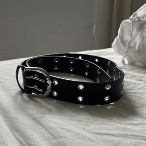 Svart bälte med silverdetaljer - Säljer ett svart bälte med dubbla rader av silverfärgade hål och ett stort silverspänne. Bältet har en cool och edgy stil, perfekt för att lyfta vilken outfit som helst.