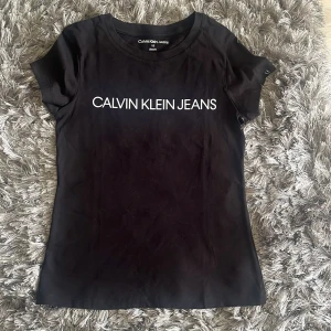 Svart t-shirt från Calvin Klein Jeans - Snygg svart t-shirt från Calvin Klein Jeans med vit logga tryckt på bröstet. Modellen är slim fit och har korta ärmar. Perfekt basplagg med stilren design och rund halsringning. Aldrig använd. 