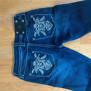 Blå jeansbyxor med broderade bakfickor från Miss Me - Säljer ett par blå jeansbyxor från Miss Me med snygga broderade detaljer på bakfickorna och vita kontrastsömmar. Byxorna har bootcut-modell och klassisk femficksdesign. Perfekta för dig som vill sticka ut lite extra!