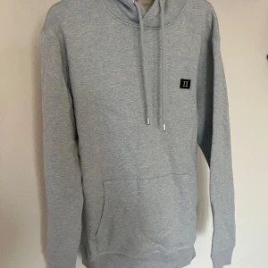 Ljusgrå hoodie från Les Deux - Säljer en stilren ljusgrå hoodie från Les Deux med broderad logga på bröstet. Tröjan har huva med dragsko och en stor magficka. Perfekt för en avslappnad look.