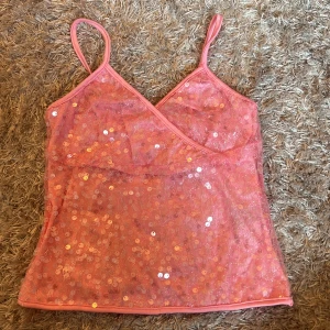 Rosa paljettlinne med smala axelband - Säljer ett glittrigt rosa linne med smala axelband och omlott-v-ringning. Toppen är täckt av paljetter som ger ett riktigt festligt intryck. Perfekt för dig som vill sticka ut och glänsa lite extra!