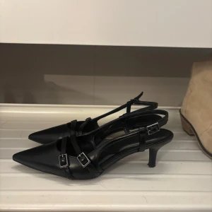 Svarta klackar med spännen - Pris kan diskuteras! Säljer ett par svarta slingback-klackar med spetsig tå och smala remmar med silvriga spännen. Skorna har öppen häl och låg smal klack. Skorna är köpta i butiken Catwalk.
