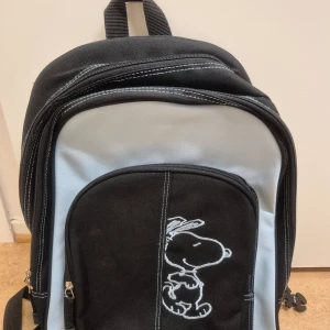 Svart och ljusblå ryggsäck med Snoopy-motiv - Säljer en svart och ljusblå ryggsäck med ett stort Snoopy-tryck på framsidan. Väskan har flera fack med dragkedja och en söt Snoopy-nyckelring. Perfekt för skolan eller vardagsäventyr!