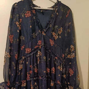 Blommig mörkblå klänning med volangdetaljer - Säljer en mörkblå klänning med blommigt mönster i rosa och beige toner. Klänningen har v-ringning, långa transparenta ärmar och flera volangdetaljer. Perfekt för vår och sommar, med en luftig och romantisk känsla. Den korrekta färgen visas på bild 1.  Dragkedja i midjan.  Storlek XL men skulle nog säga L.  Köpt på Vero Moda.  Inte använd många gånger, så i fint skick.  Den går till över knäna på mig som är 175 cm. Ej genomskinlig, finns underklänning. 