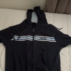 Svart hoodiejacka med dragkedja från BOSS - Svart hoodiejacka från BOSS med vit och ljusblå rand över bröstet och logga. Jackan har huva med dragsko och hel dragkedja framtill. Mjukt innerfoder och klassisk passform, perfekt för en avslappnad stil.