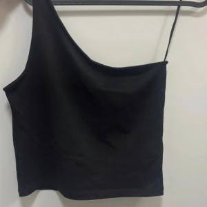 Svart oneshoulder linne - Snygg svart oneshoulder linne med smal axelrem på ena sidan. Lite ribbat material. Perfekt för att skapa en trendig och stilren look. Enkel att matcha med både jeans och kjol.