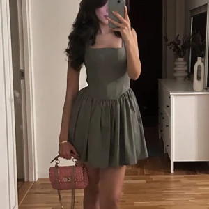 Grå kortklänning med fyrkantig urringning - Supersöt grå klänning med markerad midja och utställd kjol. Klänningen har breda axelband och en fyrkantig urringning som ger en trendig look. Perfekt för dig som vill ha en stilren men ändå lekfull outfit.