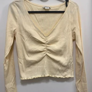 Gul långärmad topp med v-ringning från Monki - Säljer en gul långärmad topp från Monki med djup v-ringning och rynkdetalj framtill. Toppen har croppad passform och är tillverkad i ett mjukt material. Perfekt att styla med jeans eller kjol!