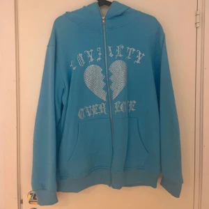 Blå kofta med strassdetaljer från Trotherz - Säljer en ljusblå kofta från Trotherz med dragkedja och huva. Framsidan har ett stort hjärta i strass och texten 'Loyalty Over Love' i glittriga bokstäver. Två fickor framtill och ribbade muddar vid ärmslut och nederkant.