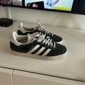 Adidas Gazelle svarta sneakers - Säljer ett par klassiska Adidas Gazelle sneakers i svart mocka med vita detaljer och vita ränder på sidorna. Skorna har vit sula och snörning framtill. Perfekta för en stilren och sportig look. Skulle behöva en tvätt så blir den fina välanvända men har mycket kvar att ge storlek 38