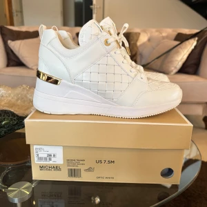 Vita sneakers med gulddetaljer från Michael Kors - Säljer ett par vita sneakers från Michael Kors med quiltad design och gulddetaljer vid hälen. Skorna har snörning och en chunky plattformssula som ger extra höjd. Perfekta för dig som vill ha en stilren och lyxig look.