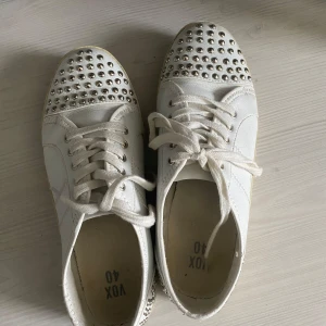 Vita sneakers med nitar från VOX - Säljer ett par vita sneakers från VOX med coola silvriga nitar framtill och baktill. Skorna har snörning och en klassisk rund tå. Perfekta för dig som vill sticka ut lite extra med din stil!