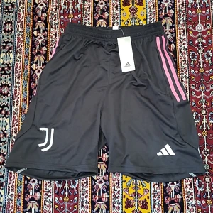 Svarta Juventus shorts från Adidas med rosa detaljer - Svarta fotbollsshorts från Adidas med rosa ränder längs sidorna och Juventus-logga på ena benet. Shortsen har snörning i midjan och dragkedjeförsedda fickor. Tillverkade i lätt och ventilerande polyester.