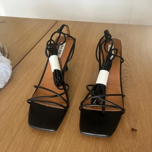 Svarta stilettklackar med snörning från NA-KD - Säljer ett par svarta stilettklackar från NA-KD med smala remmar och snörning som går att vira runt benet. Skorna har fyrkantig tå och en medelhög klack. Perfekt till alla möjliga tillfällen😍klackarna är helt oanvända, tillkommande skolåda finns med att packetera dom i💕 