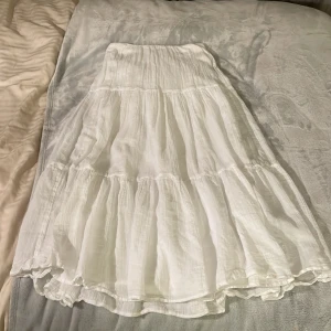 Vit volangkjol från Gina Tricot - Säljer en superfin vit långkjol från Gina Tricot med flera volanger. Kjolen har resår i midjan och är gjord i ett lätt och luftigt bomullstyg, perfekt för varma dagar. Enkel att styla med både toppar och t-shirts.