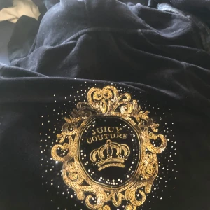 Svart velourjacka från Juicy Couture - Juicy Couture svart velourjacka med ikoniskt guldfärgat broderi och krona på ryggen. Jackan har dragkedja framtill och är långärmad. Perfekt för en trendig och avslappnad stil.