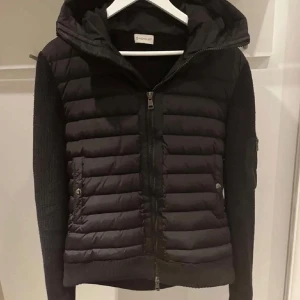 Moncler cardigan  - Säljer en svart pufferjacka från Moncler med stickade ärmar och huva. Jackan har dragkedja framtill, två fickor med knappar och Moncler-logga på ärmen. Perfekt för kyliga dagar och snygg till vardags. Aldrig använd 10/10 skick , säljer för den passade liten