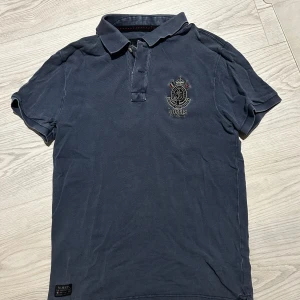 Mörkblå pikétröja från Morris - Säljer en mörkblå pikétröja från Morris med klassisk krage och korta ärmar. Tröjan har broderad logga på bröstet och knäppning vid halsen. Perfekt för en stilren och avslappnad look. Tröjan är hel och fin, dock något tvettblekt, framförallt runt krage, ärmar och sömmar. 