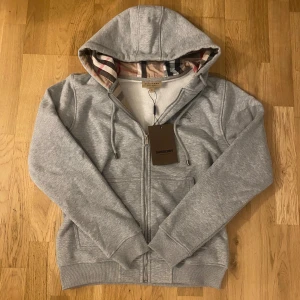 Grå hoodie från Burberry - Säljer en snygg grå hoodie från Burberry med dragkedja och klassiskt rutigt foder i huvan. Tröjan har långa ärmar, fickor framtill och justerbar huva med snören. Perfekt för en stilren och bekväm look.