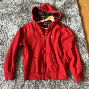 Röd hoodie från Polo Ralph Lauren - Röd hoodie från Polo Ralph Lauren med dragkedja och huva. Klassisk gul logga broderad på bröstet och rutigt innerfoder i huvan. Två fickor framtill och ribbade muddar vid ärmslut och nederkant. Skön inför vår och sommaren. Pris går att diskutera vid snabb affär
