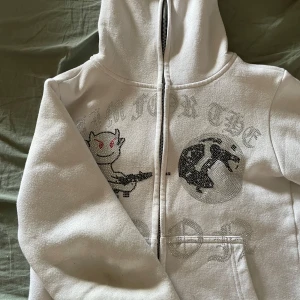 Vit hoodie med glittrigt tryck och djävulsmotiv - Vit hoodie med dragkedja och huva. Framsidan har ett glittrigt tryck med en tecknad djävul som håller i ett vapen samt en jordglob. Text i silver och stenar. Perfekt för dig som gillar unika och lekfulla plagg.