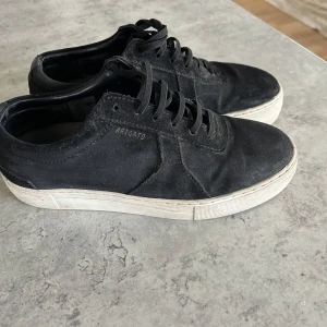 Svarta sneakers från Axel Arigato - Snygga svarta sneakers från Axel Arigato med ovandel i mocka och klassisk vit platt sula. Skorna har snörning och diskret guldfärgad logga på sidan. Perfekta för en stilren och modern look. Använda en säsong sparsamt men syns såklart på sula och lite på mockan. Väl omhändertagna. Ordniarie pris 2550kr 