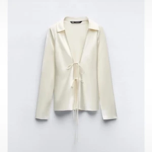 Vit satinblus med knytning från Zara - Snygg vit blus i satin från Zara med lång ärm och djup v-ringning. Blusen har knytning framtill och en klassisk krage. Perfekt för att skapa en stilren och trendig look.