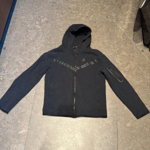 Svart  Nikehoodie stl 158-170 - Svart hoodie från Nike med dragkedja framtill och huva. Jackan har diskreta svarta detaljer på bröstet och en ficka med dragkedja på ena ärmen. Liten defekt vid ena ärmslutet (se sista bilden)