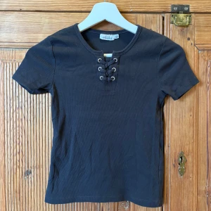 Mörkgrå ribbad topp med snörning från KappAhl - Mörkgrå ribbad topp från KappAhl med korta ärmar och snygg snörningsdetalj vid halsen. Perfekt för en stilren och trendig look. Mjuk och bekväm att bära. Storlek 146-152.