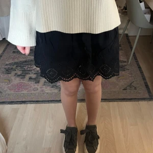 Svart kjol med broderade detaljer - Säljer en svart kortkjol med bred resår i midjan och fina broderade håldetaljer längs fållen. Kjolen har en lätt utställd form och är perfekt för dig som gillar en enkel men ändå detaljrik stil.