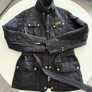 Svart quiltad jacka från Barbour med bälte - Säljer en svart quiltad jacka från Barbour med guldiga detaljer, bälte i midjan och hög krage. Jackan har flera fickor med knappar och dragkedja framtill. Perfekt för dig som vill ha en klassisk och stilren look.