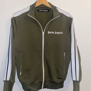 Olivgrön tröja från Palm Angels - Säljer en olivgrön track jacket från Palm Angels med vita ränder längs ärmarna och vit dragkedja. Jackan har hög krage, två fickor med dragkedja och logga på bröstet. Perfekt för en sportig look.Tröjan är en i en bra skick. Säljer den för var liten på mig när den kom.
