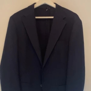 Mörkblå ullkavaj från These Glory Days - Säljer en mörkblå kavaj från These Glory Days i 100% ull (Super 100's Wool). Klassisk passform med två knappar framtill och fyra dekorativa knappar vid ärmslutet. Perfekt för bal eller fest. Knappt använd och passar dig som är runt 185cm.