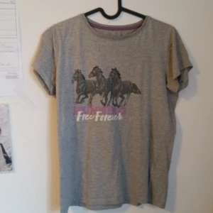 Grå t-shirt med hästmotiv och text - Grå t-shirt med tryck av tre svarta hästar och texten 'Free Forever' i vitt och rosa. Rund halsringning och korta ärmar. Perfekt för dig som gillar hästar och vill ha en avslappnad stil.