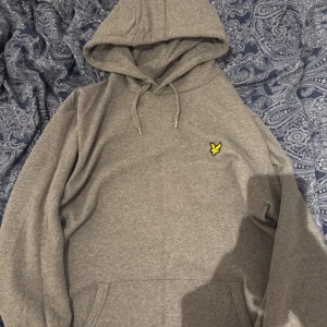 Grå hoodie från Lyle & Scott - Säljer en grå hoodie från Lyle & Scott med klassisk gul logga på bröstet. Tröjan har huva med dragsko och en stor magficka. Tillverkad i ekologisk bomull för extra komfort. Tröjan är storlek L men passar M också. 