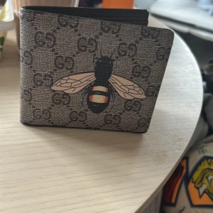 Gucci plånbok med bi-motiv - Säljer en snygg plånbok från Gucci med klassiskt GG-mönster och en stor broderad bi på framsidan. Plånboken är grå med svarta och beige detaljer och har flera fack för kort och sedlar. Perfekt accessoar för dig som gillar designer.